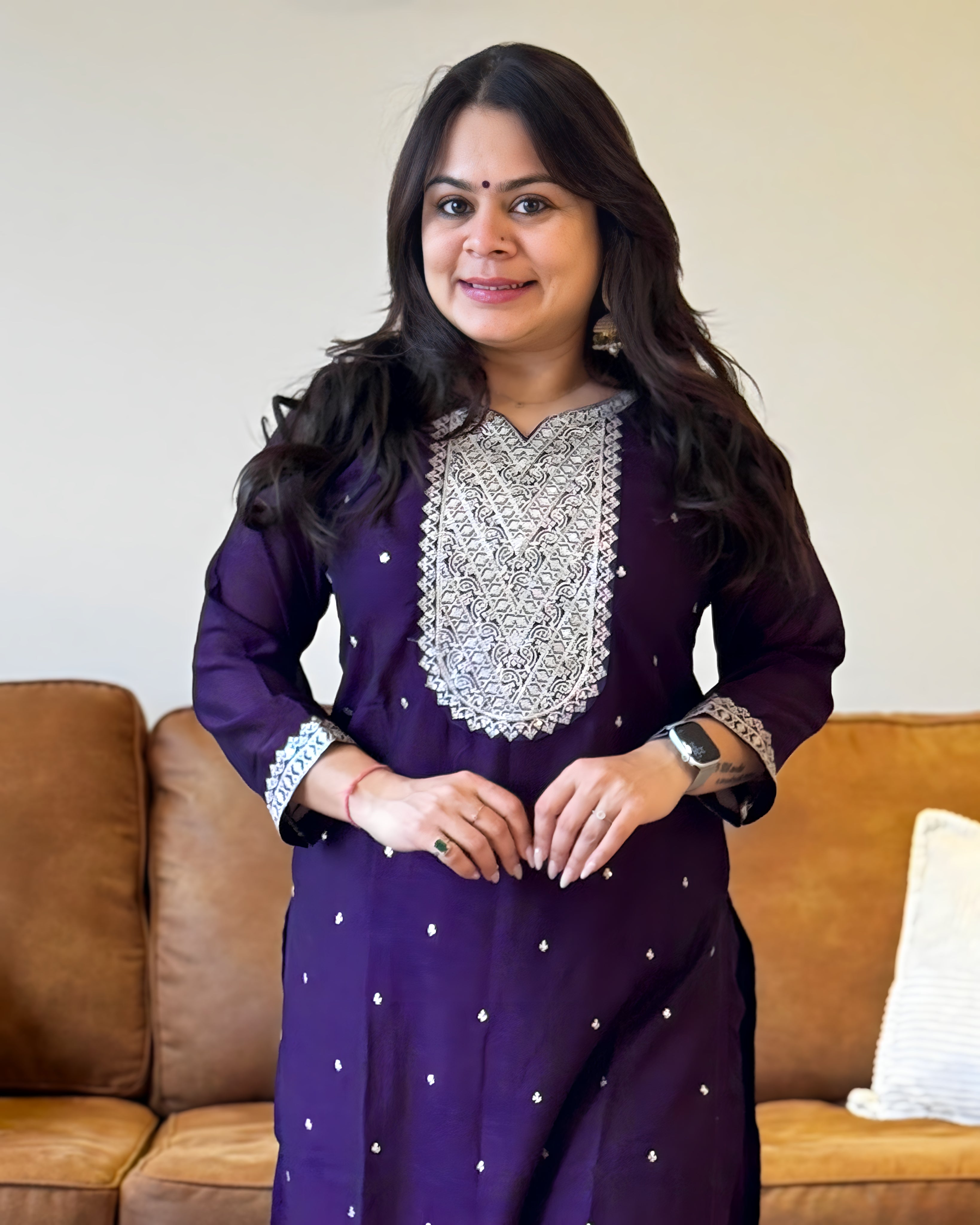 Woman wearing a purple traditional outfit with silver embroidery in a living room setting.