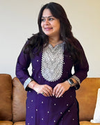 Woman wearing a purple traditional outfit with silver embroidery in a living room setting.