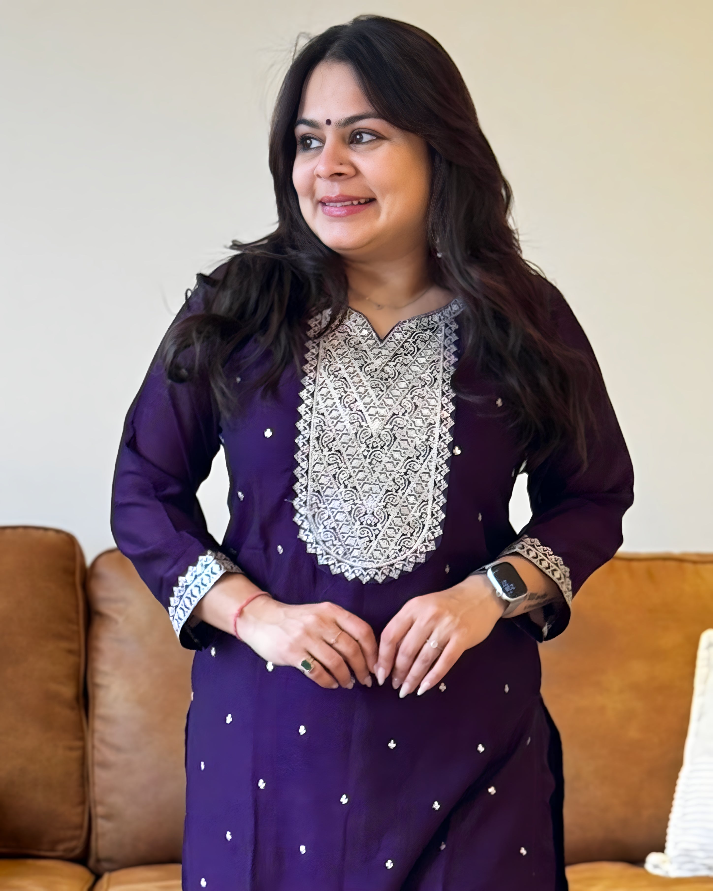 Woman wearing a purple traditional outfit with silver embroidery in a living room setting.