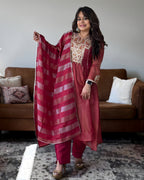 A woman models a traditional Indian salwar kameez set in a living room setting. The outfit consists of a dusty rose pink tunic with gold embroidery, matching pink trousers, and a striped dupatta in shades of pink and red with gold borders.