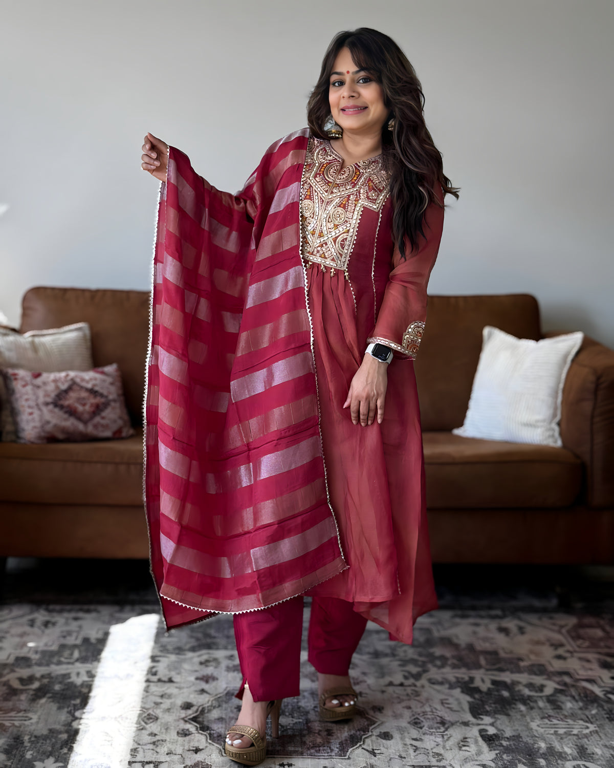 A woman models a traditional Indian salwar kameez set in a living room setting. The outfit consists of a dusty rose pink tunic with gold embroidery, matching pink trousers, and a striped dupatta in shades of pink and red with gold borders.