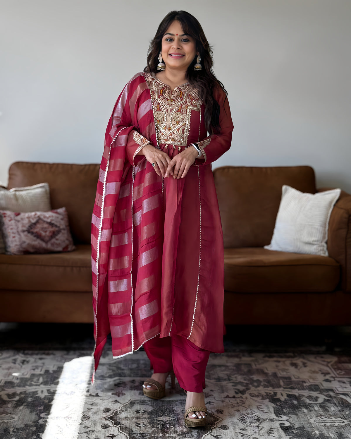 A woman modeling a traditional Indian salwar kameez suit in a living room setting. The outfit consists of a deep magenta-pink kurta with intricate gold embroidery on the chest, matching pink trousers, and a striped dupatta with gold borders.