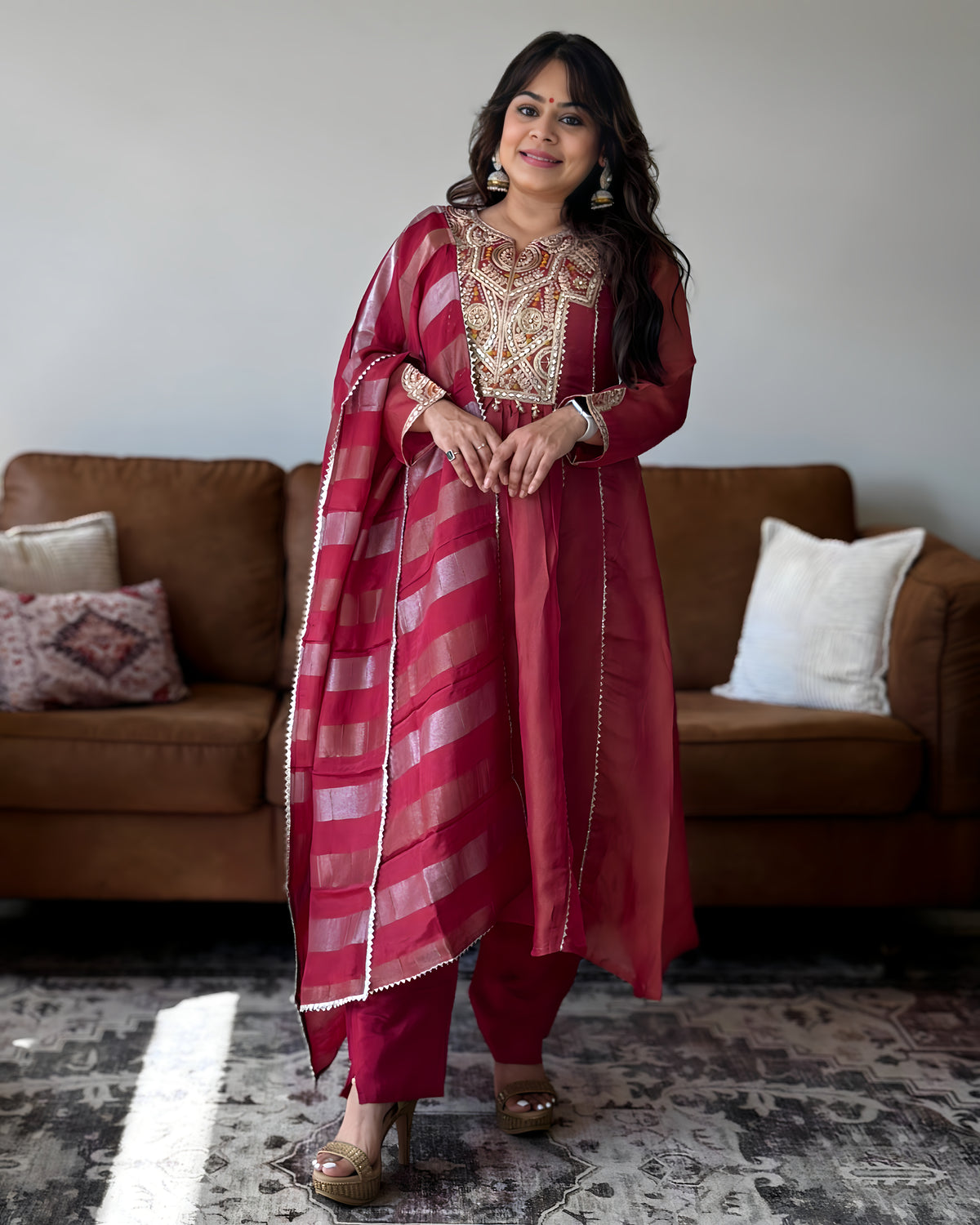 A woman models a traditional Indian salwar kameez suit in a living room setting. The outfit consists of a deep pinkish-red kurta with gold embroidery on the chest, matching trousers, and a striped dupatta in shades of red and pink with gold borders.