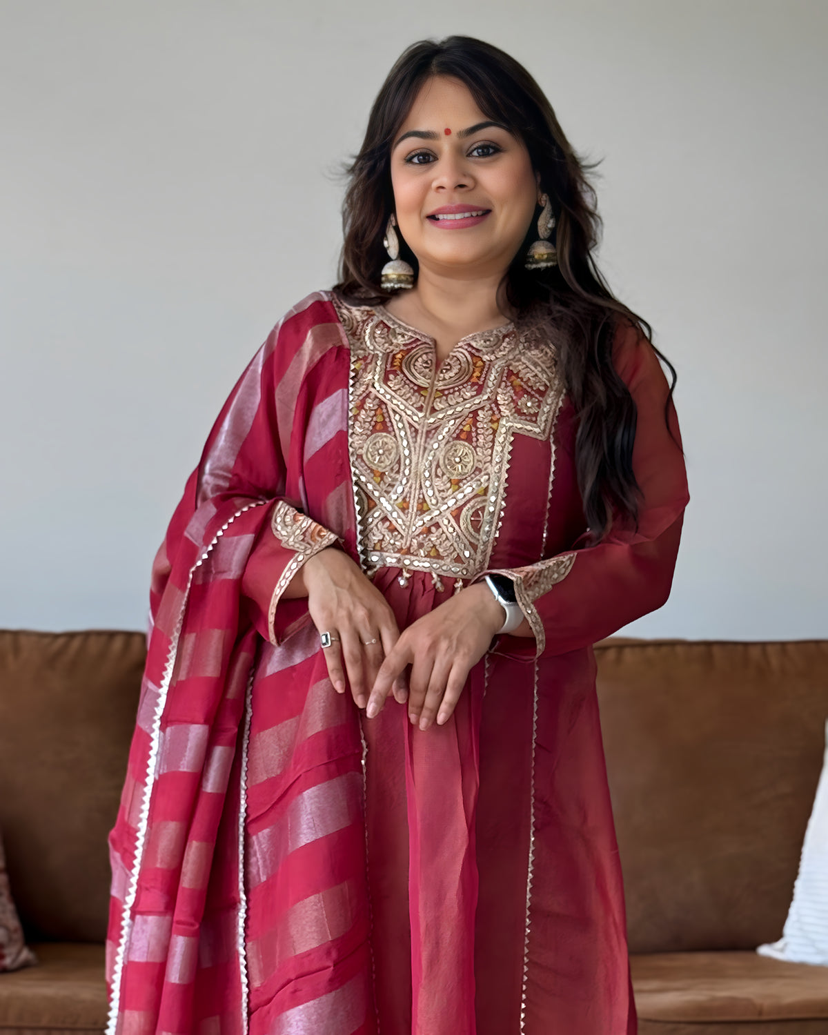 A woman wearing a traditional Indian salwar kameez outfit in deep red and pink with intricate gold embroidery, standing in front of a neutral wall and a brown sofa.
