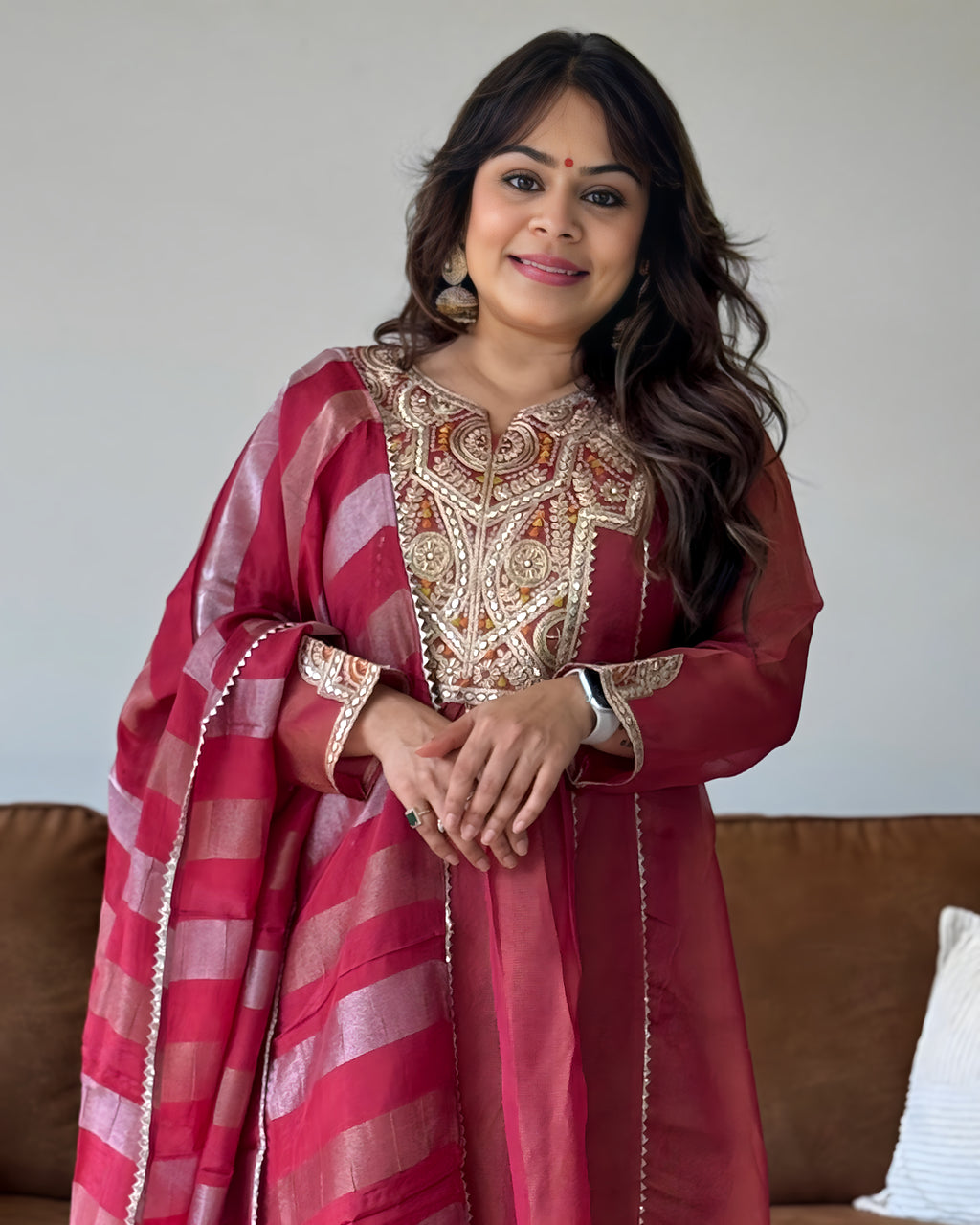 A woman wearing a traditional Indian outfit consisting of a deep red kurta with intricate gold embroidery and a matching striped dupatta, standing in front of a neutral wall and a brown sofa.