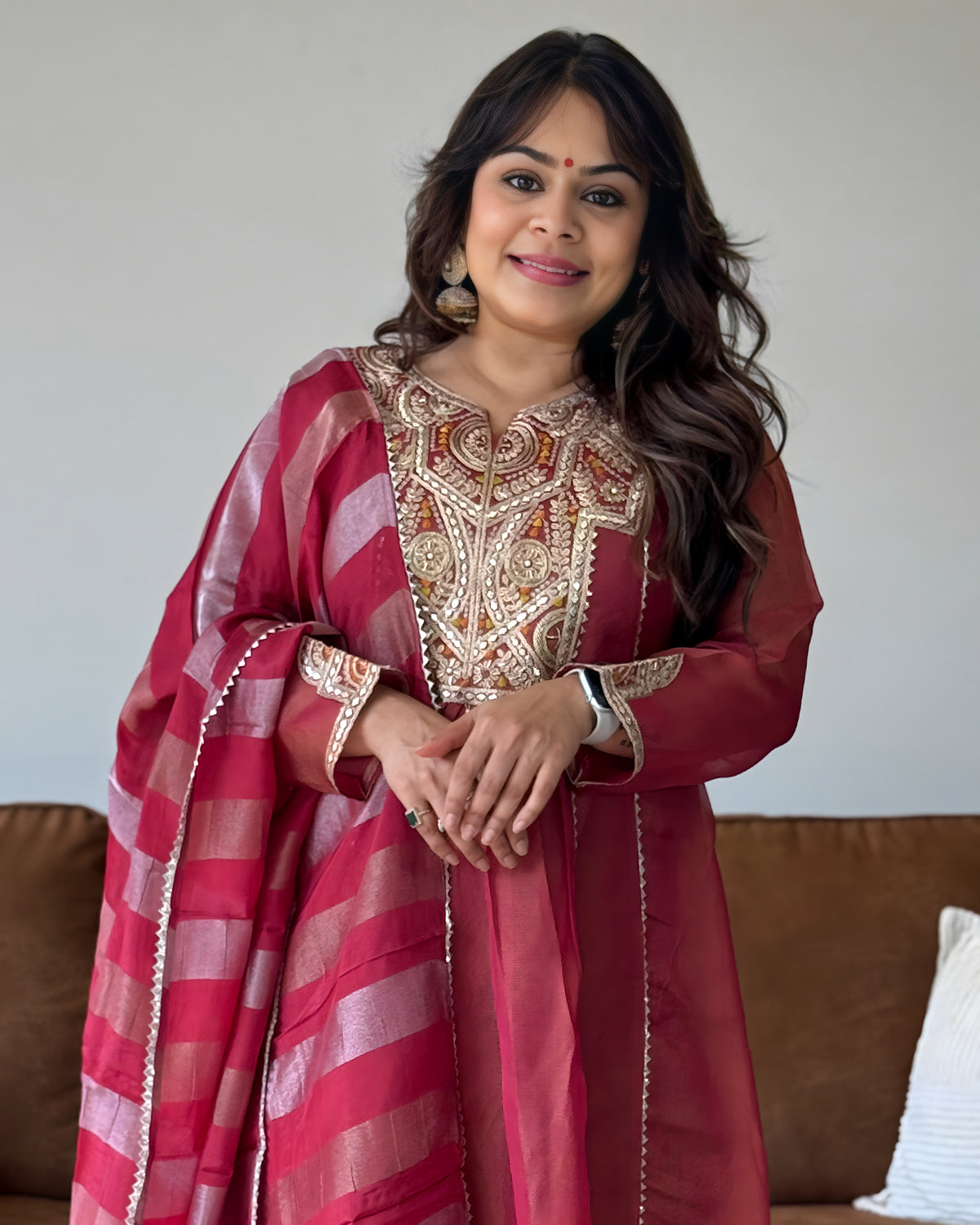 A woman wearing a traditional Indian outfit consisting of a deep red kurta with intricate gold embroidery and a matching striped dupatta, standing in front of a neutral wall and a brown sofa.
