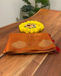 Orange fabric with gold patterns and red tassels on a wooden surface with yellow flowers and a decorative item.