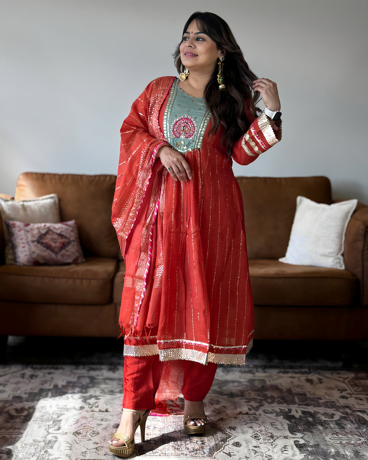 A woman models a traditional Indian salwar kameez suit in a living room setting. The outfit consists of a coral-red kurta with gold zari embroidery, a light teal-green inner lining with a red and gold circular mandarin, matching red trousers, and a coral-red dupatta with gold patterns.