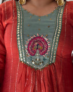 Close-up of a traditional Indian outfit featuring a coral red kurta with a teal embroidered yoke. The chest area is adorned with a detailed peacock motif in pink, green, and gold, surrounded by gold coin embellishments and vertical silver bead lines.