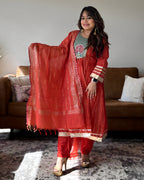 A woman models a red Indian ethnic suit set featuring a kurta, trousers, and a dupatta with gold zari embroidery, standing in a living room setting.