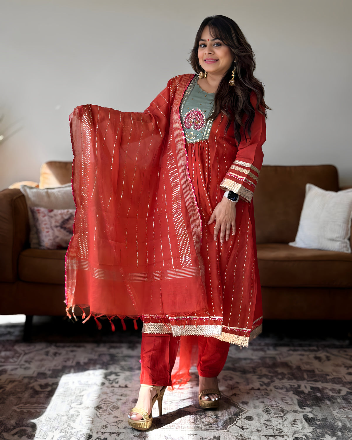 A woman models a red Indian ethnic suit set featuring a kurta, trousers, and a dupatta with gold zari embroidery, standing in a living room setting.