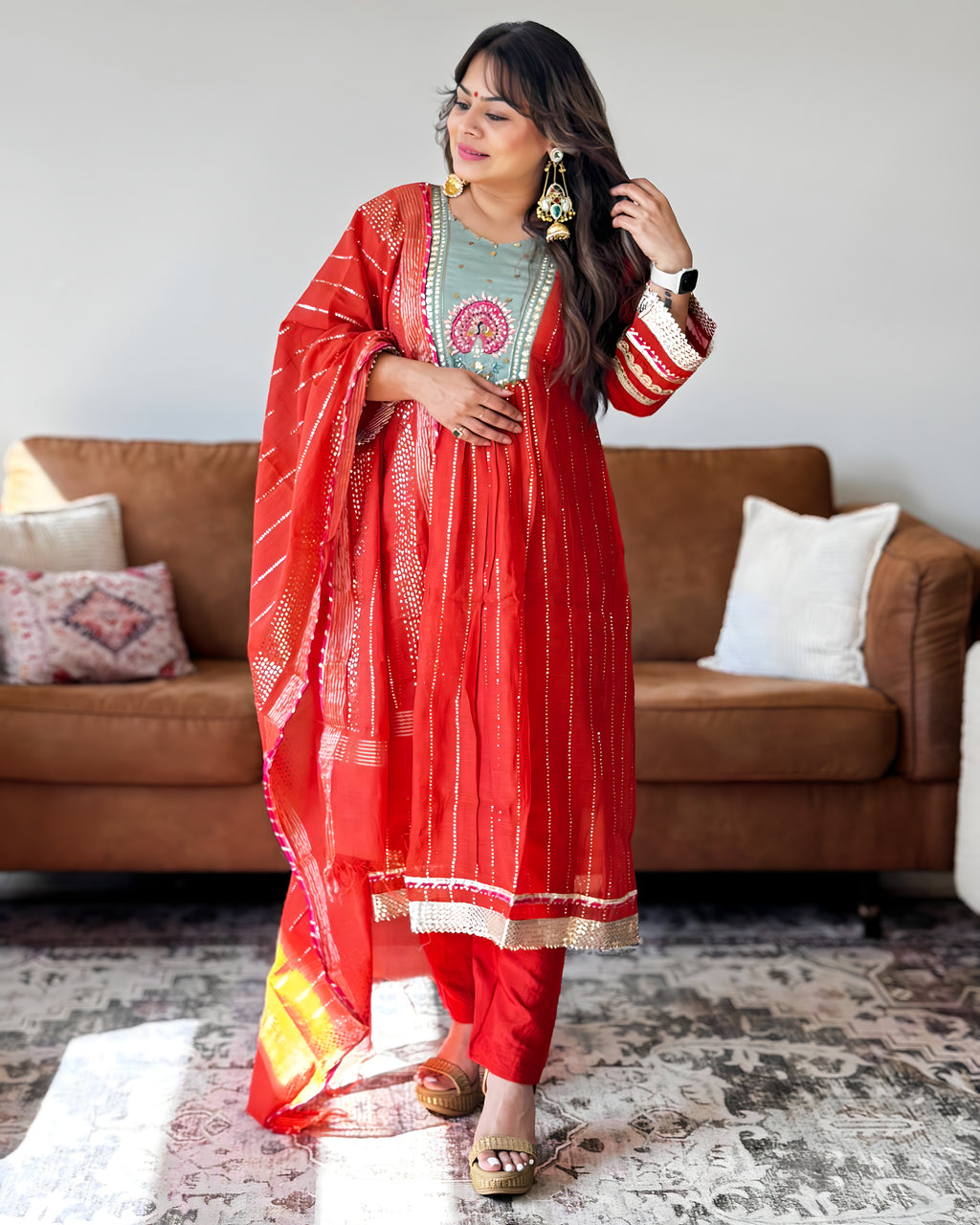 A woman models a traditional Indian ethnic outfit consisting of a red kurta with gold zari embroidery, matching red pants, and a red dupatta, set against a living room background with a brown sofa.