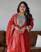 A woman wearing a traditional red and gold embroidered Anarkali kurta with a light blue-green embroidered neckline, standing in front of a brown sofa.