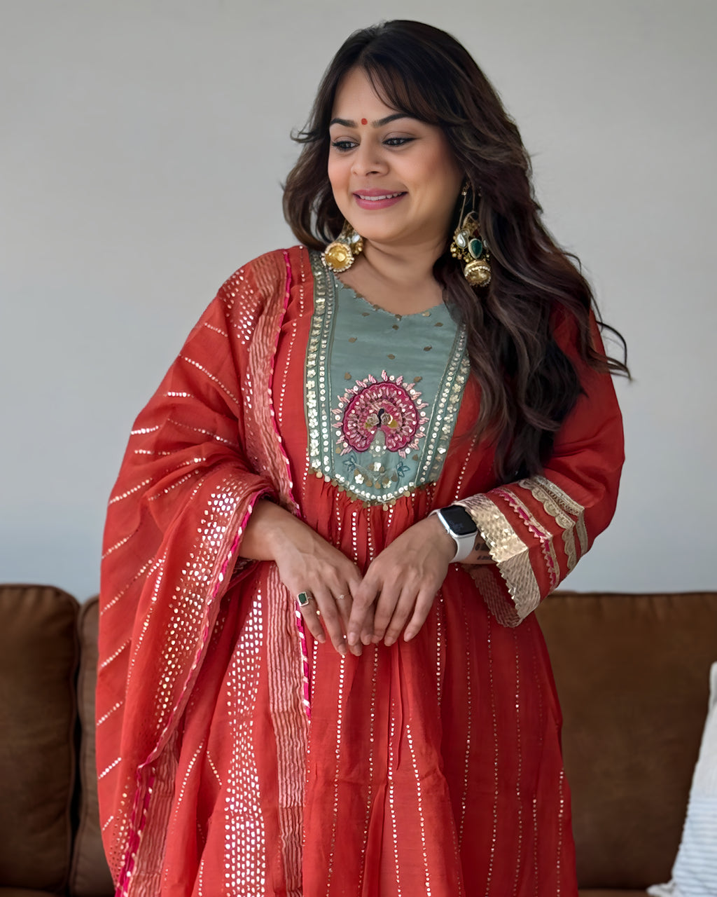 A woman wearing a traditional Indian outfit consisting of a coral red kurta with gold zari embroidery, a light sage green embroidered yoke, and a matching coral red dupatta draped over her shoulders. She is also wearing large gold earrings and a smartwatch.