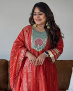 A woman wearing a traditional Indian outfit consisting of a coral red kurta with gold zari embroidery, a light sage green embroidered yoke, and a matching coral red dupatta draped over her shoulders. She is also wearing large gold earrings and a smartwatch.