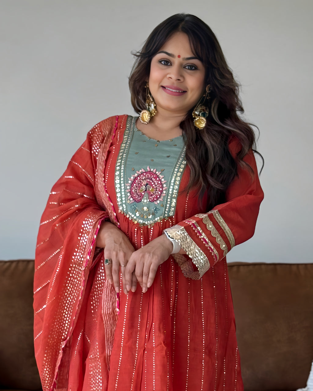 A woman wearing a traditional Indian outfit consisting of a coral red anarkali kurta with gold zari embroidery, a light teal-green embroidered yoke, and a matching coral dupatta, standing in front of a neutral wall and a brown sofa.