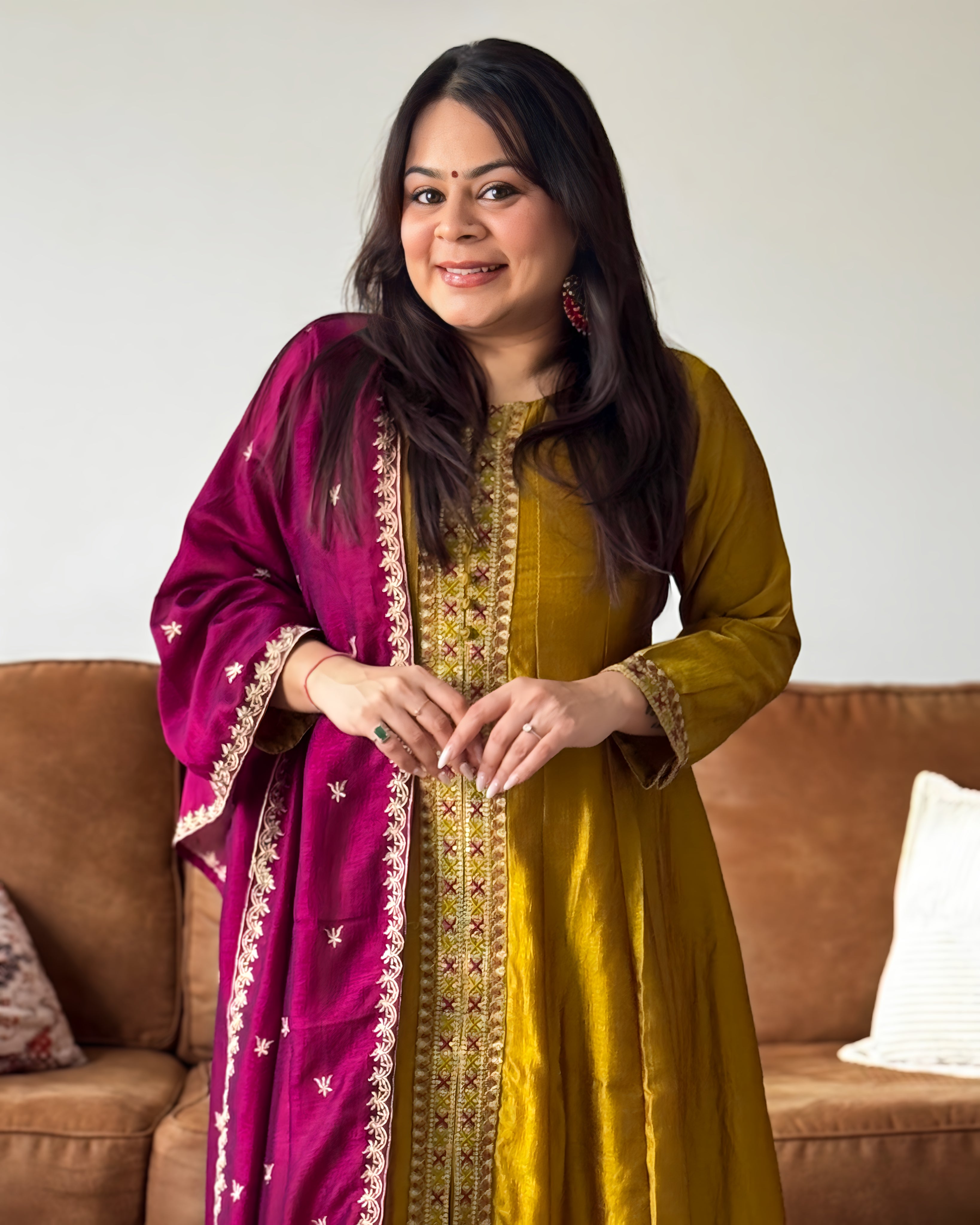 Woman wearing a yellow traditional outfit with a pink dupatta, standing in a room with a brown couch.