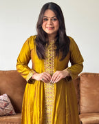 Woman wearing a yellow traditional outfit with intricate patterns, sitting on a brown couch.