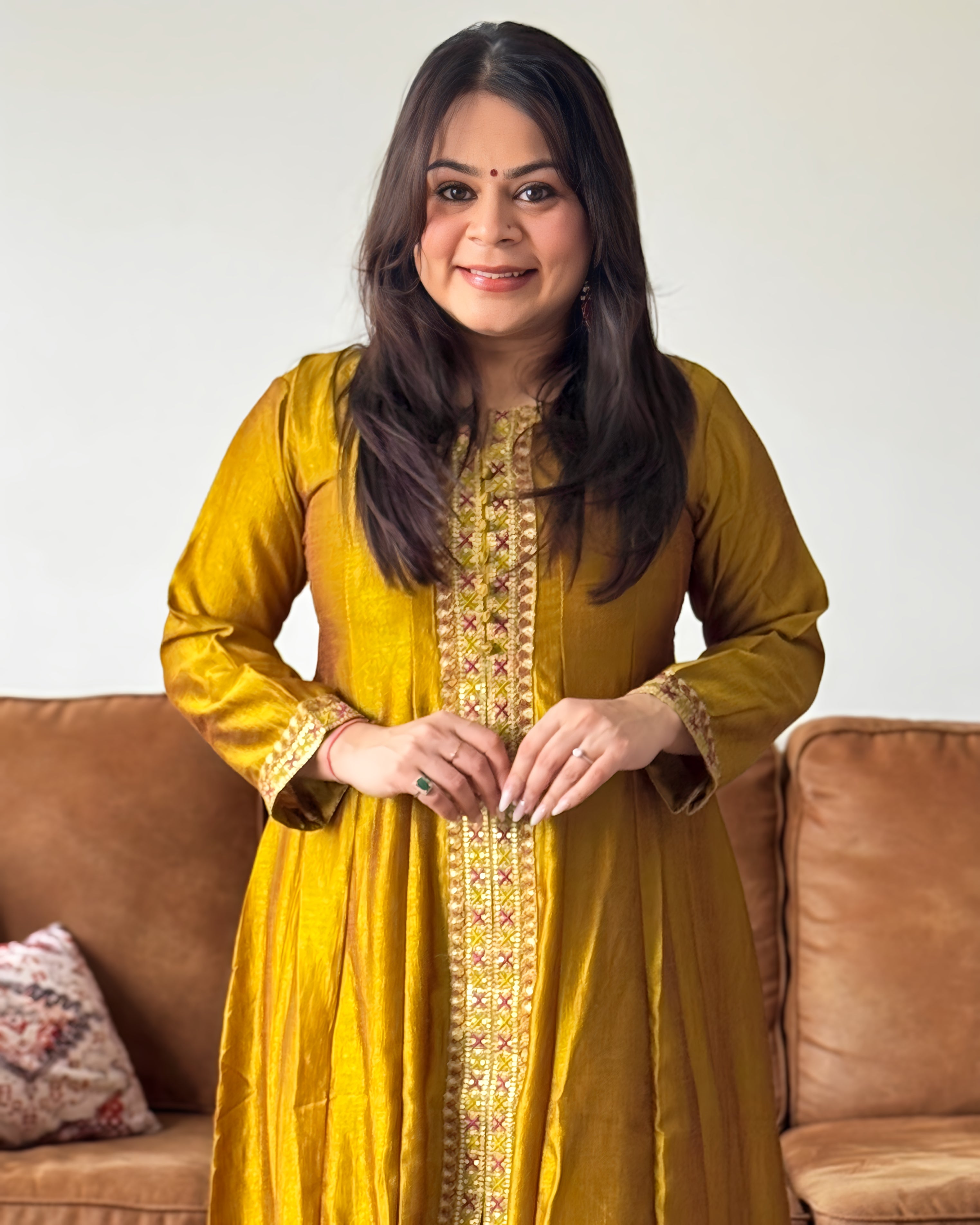 Woman wearing a yellow traditional outfit with intricate patterns, sitting on a brown couch.