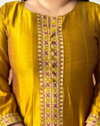 Woman in a green traditional outfit standing in a living room with a brown sofa and patterned rug.