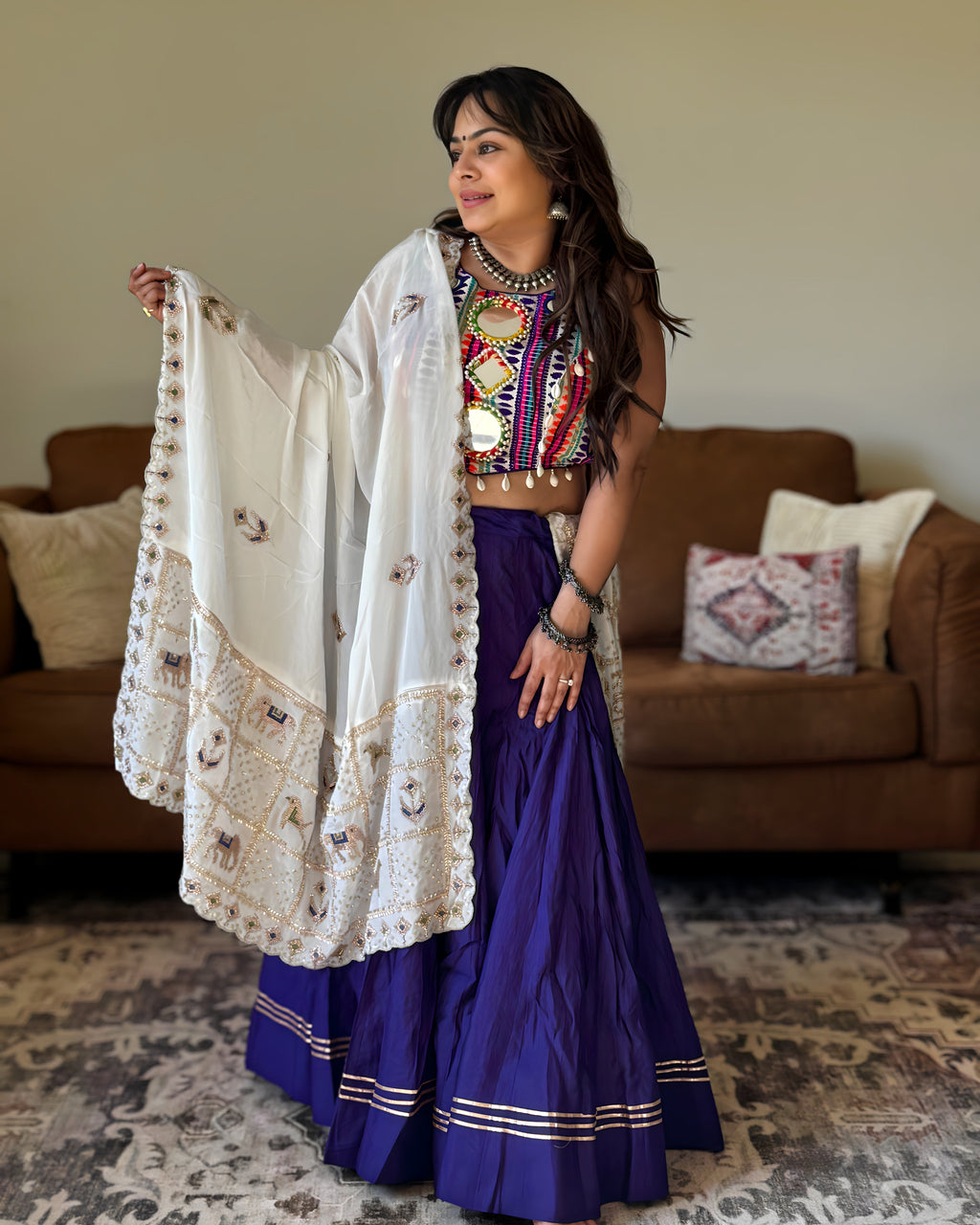 A woman models a traditional Indian outfit consisting of a deep purple skirt, a colorful embroidered crop top, and a white embroidered dupatta, standing in a living room with a brown sofa in the background.