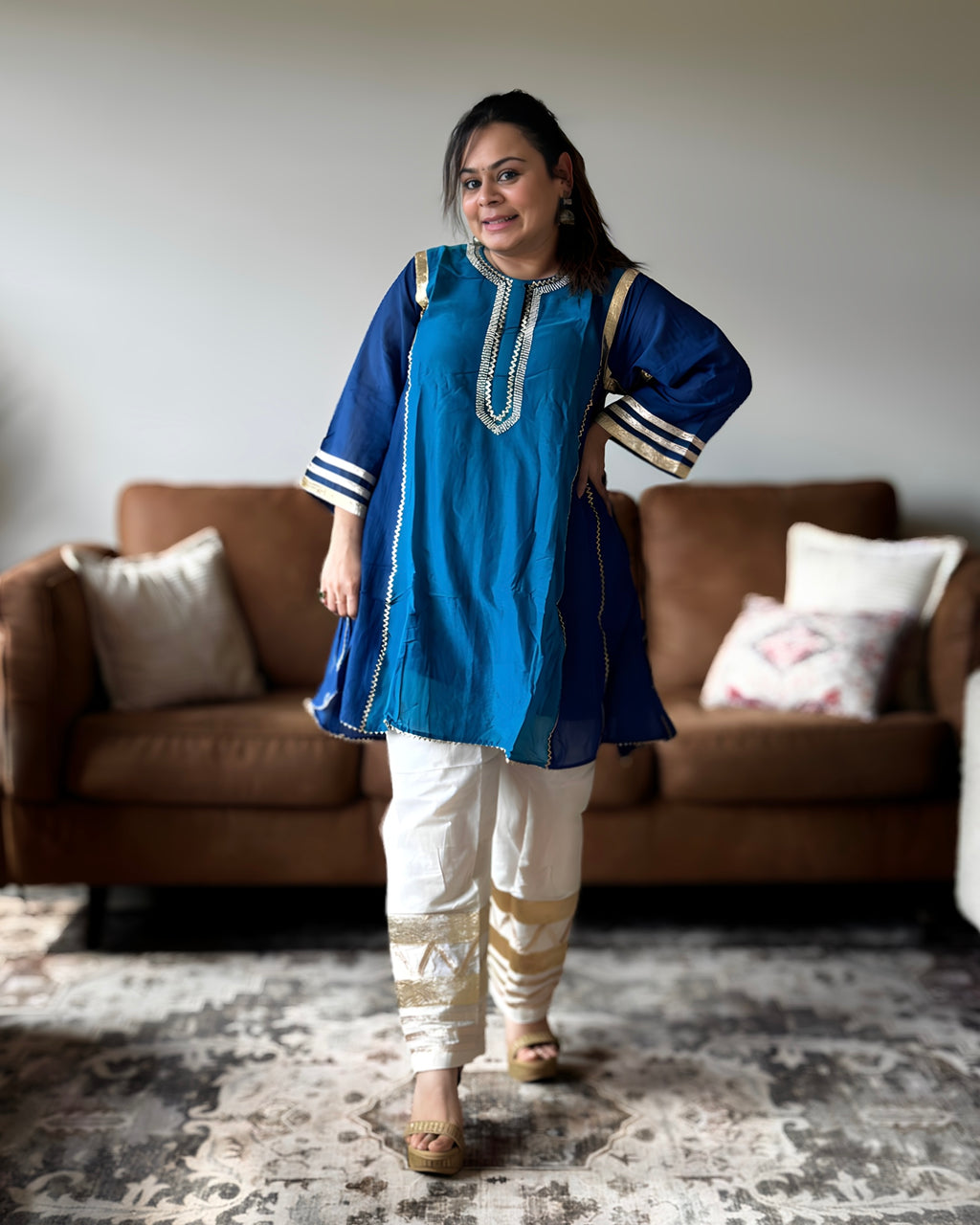 A woman modeling a blue kurta with white pants in a living room setting with a brown sofa and patterned rug.