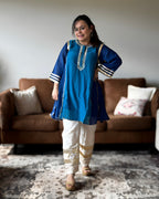 A woman modeling a blue kurta with white pants in a living room setting with a brown sofa and patterned rug.