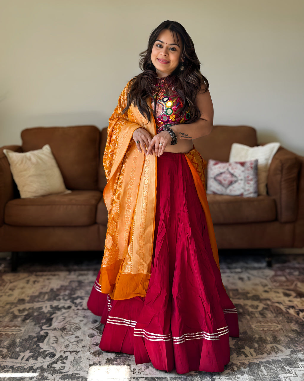 A woman wearing a traditional Indian outfit consisting of a deep red skirt, a multicolor embroidered top, and an orange dupatta, standing in a living room with a brown sofa in the background.