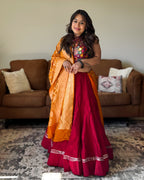 A woman wearing a traditional Indian outfit consisting of a deep red skirt, a multicolor embroidered top, and an orange dupatta, standing in a living room with a brown sofa in the background.